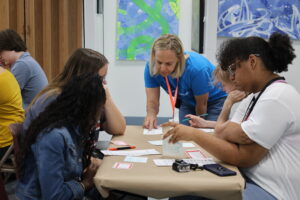 A group of individuals gathers around a table, engaged in a collaborative activity guided by a facilitator who is pointing to materials on the table.