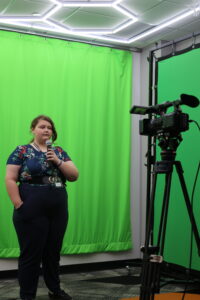 A person stands in front of a green screen holding a microphone, being recorded by a professional camera on a tripod.
