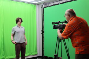 A person stands in front of a green screen while another operates a camera on a tripod.