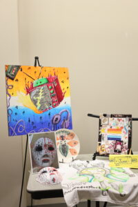 A display of colorful art pieces including a painting of a fiery television, masks, a decorated plate, a collage, and a t-shirt with a drawing, arranged on a table.
