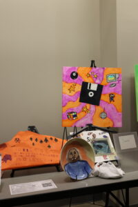 A display table features a colorful painting with retro tech items, an orange fabric artwork, a ceramic bowl with a portrait, and other creative items.