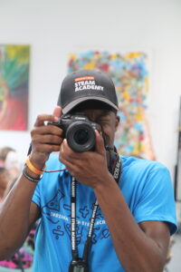 A person wearing an Indiana Tech Steam Academy cap and shirt holds a Canon EOS R6 camera to their eye, capturing a moment in a colorful indoor setting.