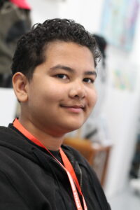 A young person with curly dark hair smiles gently while wearing a black hoodie and an orange lanyard with 