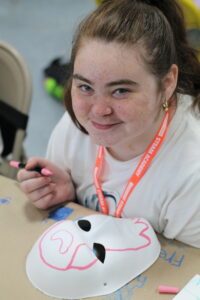 A person with freckles smiles while drawing on a white mask with a pink marker at a table.