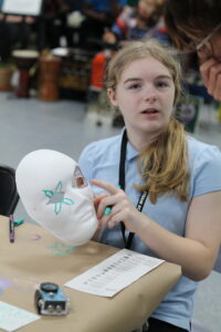 A young person holds up a white mask with teal and gray designs while pointing at it, seated at a table with art supplies and a small device.