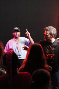 Two individuals stand on a stage, one gesturing upward while the other looks on, with an audience seated in front.
