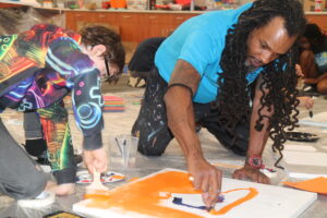 Two individuals are actively engaged in painting on a canvas, with one using a brush and the other applying paint with their fingers.