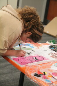 A person with curly hair leans over a table, carefully coloring a cartoon character on a large sheet of paper with a marker.