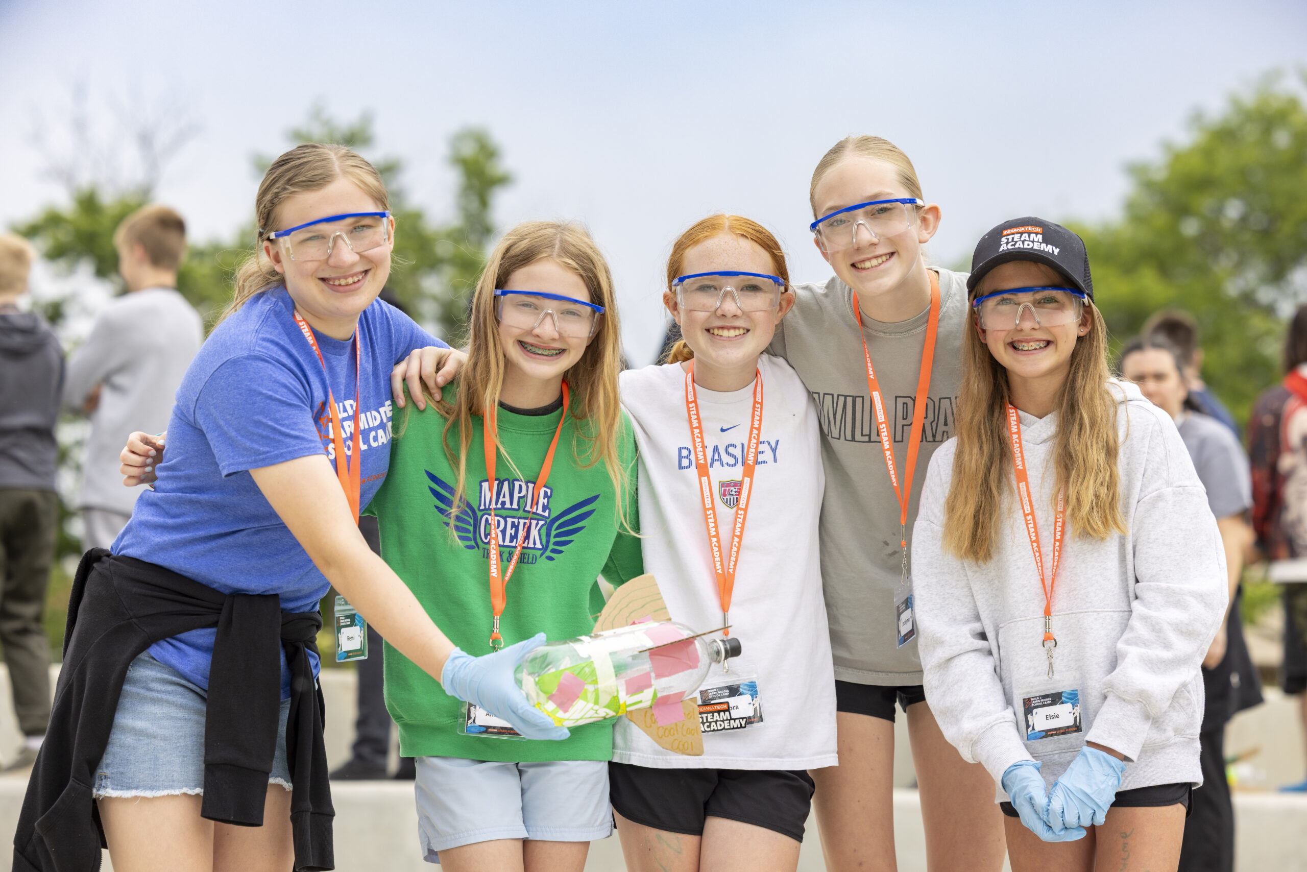 Five young individuals wearing safety goggles and gloves pose together outdoors, one holding a decorated bottle, while others wear lanyards and team apparel.
