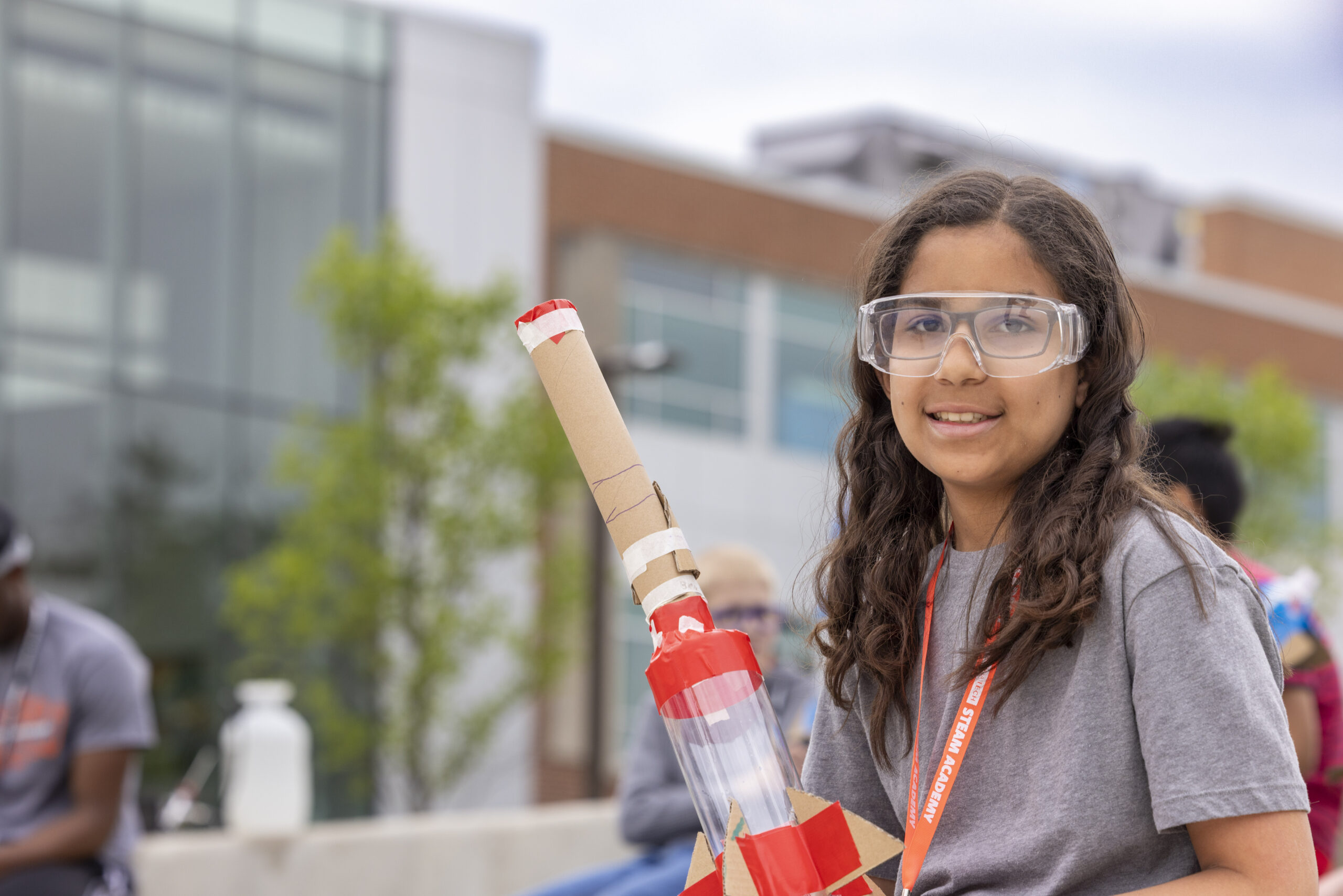 A young person wearing safety goggles smiles while holding a handmade cardboard rocket outdoors.