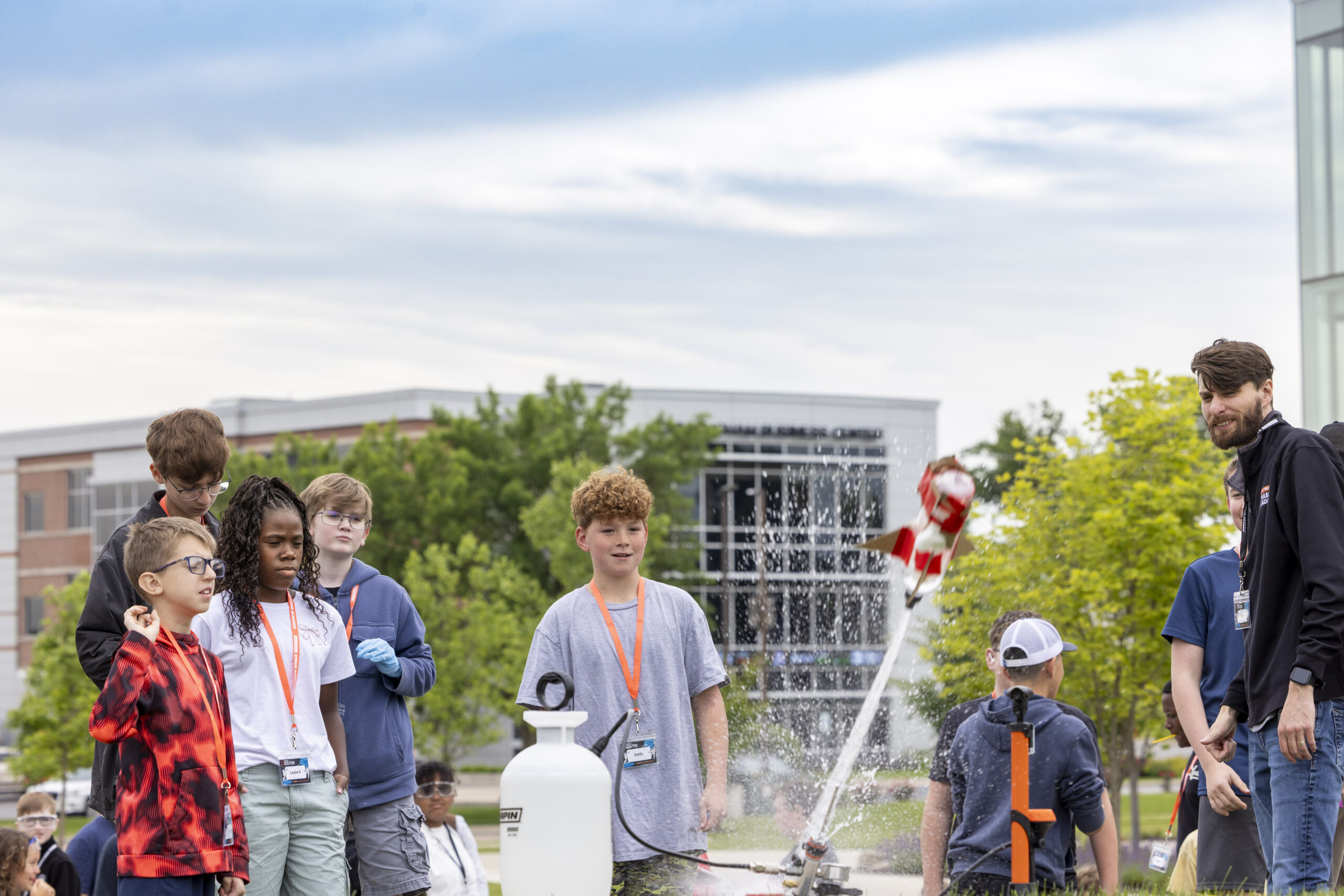 A group of children and adults observe a rocket launching from a water cannon outdoors on a cloudy day.