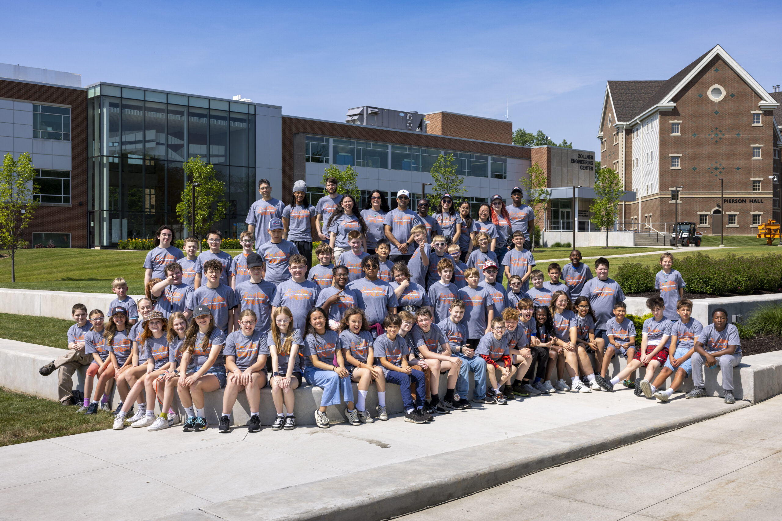 A group of young people in matching gray shirts pose outdoors in front of modern and traditional campus buildings on a sunny day.