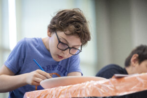 A young person with glasses carefully draws on a white plate while seated at a table.