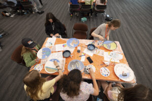 A group of children sits around a table, engaged in painting decorative plates with colorful designs.