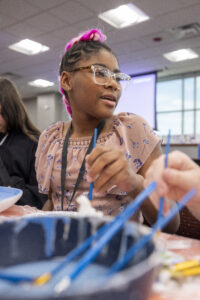 A young person with pink hair and glasses is engaged in a creative activity, holding paintbrushes and working on a project at a table.