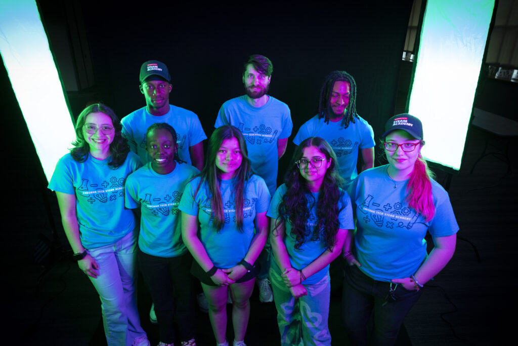A group of seven individuals wearing matching t-shirts and caps pose together under vibrant neon lighting.