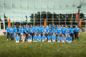A group of people wearing matching blue shirts poses for a photo on a grassy area in front of a modern glass building.
