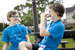 Two young individuals in matching blue shirts sit at a picnic table outdoors, smiling and interacting while one raises their fist.