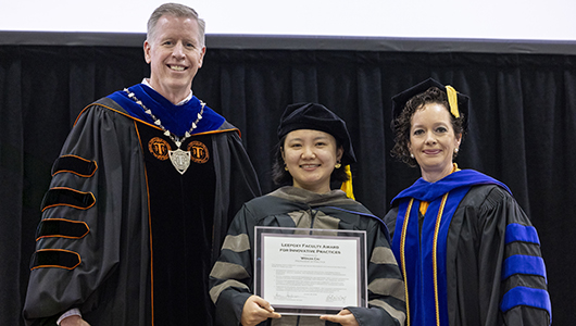 Dr. Cai, middle, holds a framed award while standing with two university officials in academic regalia.